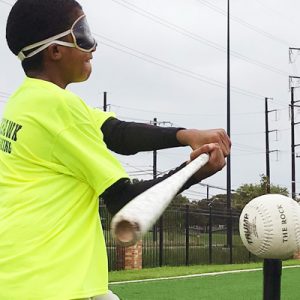 A blindfolded youth athlete nearly makes contact as she swings a bat at a ball on a tee.